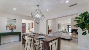 Uma área de refeições com uma mesa de madeira, cadeiras e um candelabro. Há um sofá e uma mesa de centro ao fundo.