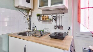 Une cuisine avec un plan de travail en bois, un évier en acier inoxydable et des armoires blanches. On y trouve une fenêtre avec un rideau rouge, un porte-ustensiles suspendu et une petite plante accrochée au mur.