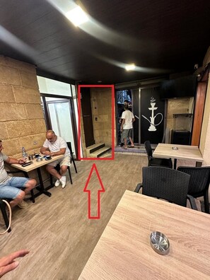 A restaurant interior with wooden tables, a black wall with a hookah symbol, and a person walking towards the door. - Timberwood Studio in Batroun (Batroun)