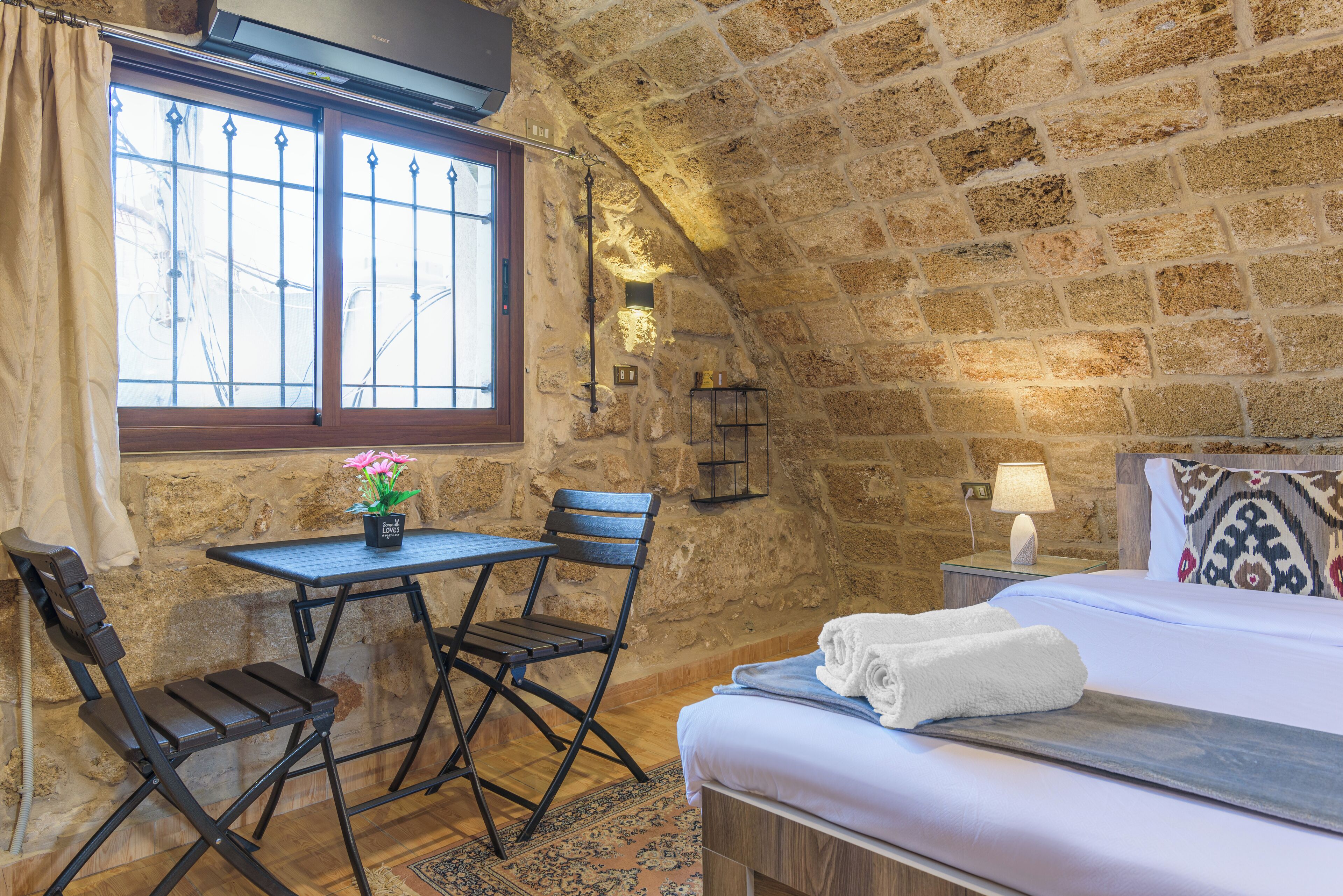 Una habitación acogedora con pared de piedra, cama con sábanas blancas, una mesita con una silla y una ventana con cortinas.