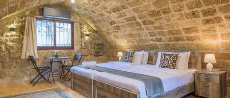 A bedroom with a stone ceiling, a wooden bed, a nightstand, a table with chairs, and a window with curtains.
