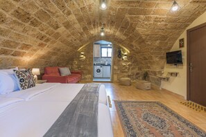 A bedroom with a stone ceiling, a bed with white linens, a red sofa, and a view into the kitchen. - Timberwood Studio in Batroun (Batroun)