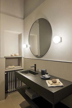 Un baño moderno con encimera oscura, un espejo redondo y un estante empotrado con artículos de aseo.