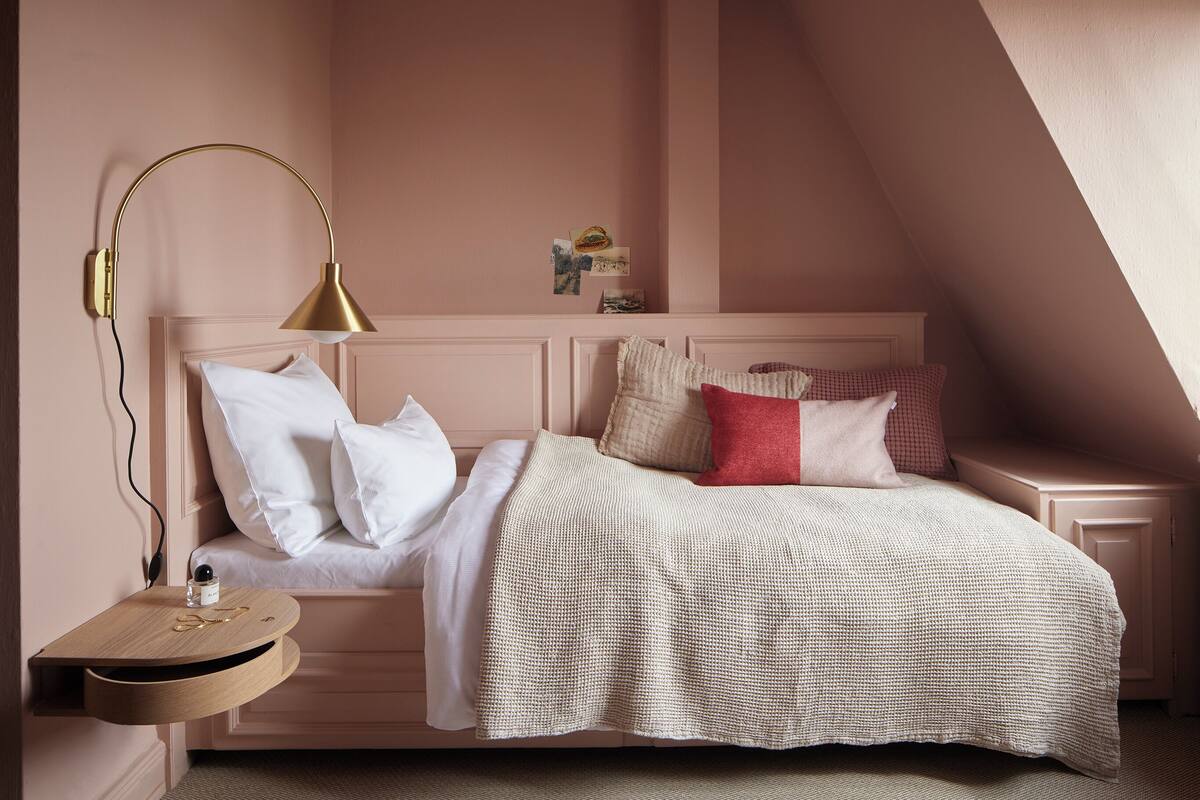 Een slaapkamer met een schuine plafond, een bed met witte en rode kussens, een nachtkastje met een lamp en een wandplank met foto’s.