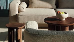 Un salon avec un canapé beige, deux petites tables en bois et un saladier de fruits posé sur l’une des tables.