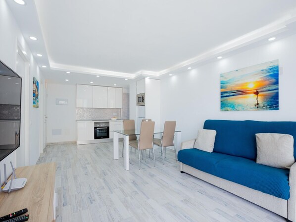 Una sala de estar moderna con un sofá azul, un comedor con una mesa de vidrio y una cocina con armarios blancos.