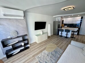 Un salon moderne doté d’un téléviseur à écran plat, d’un meuble de rangement intégré pour les appareils multimédias, d’une table basse ronde, d’un canapé et d’un coin cuisine avec un bar et des tabourets.