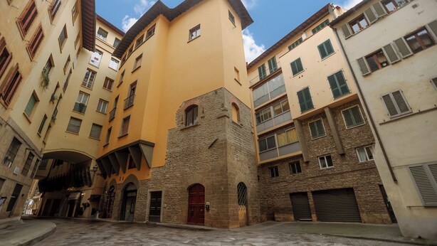 Une rue étroite, bordée de bâtiments de chaque côté, caractérisée par des façades en pierre et en stuc, des portes en arc de cercle et de nombreuses fenêtres.