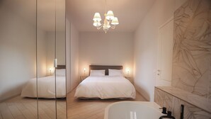 Une chambre moderne dotée d’un grand lit, d’un coin salon séparé, d’une salle de bain avec baignoire et d’un miroir qui reflète la pièce.