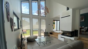 A spacious living room with a large grey sofa, a glass coffee table, and a TV. There are large windows providing natural light and a view of the outdoors.