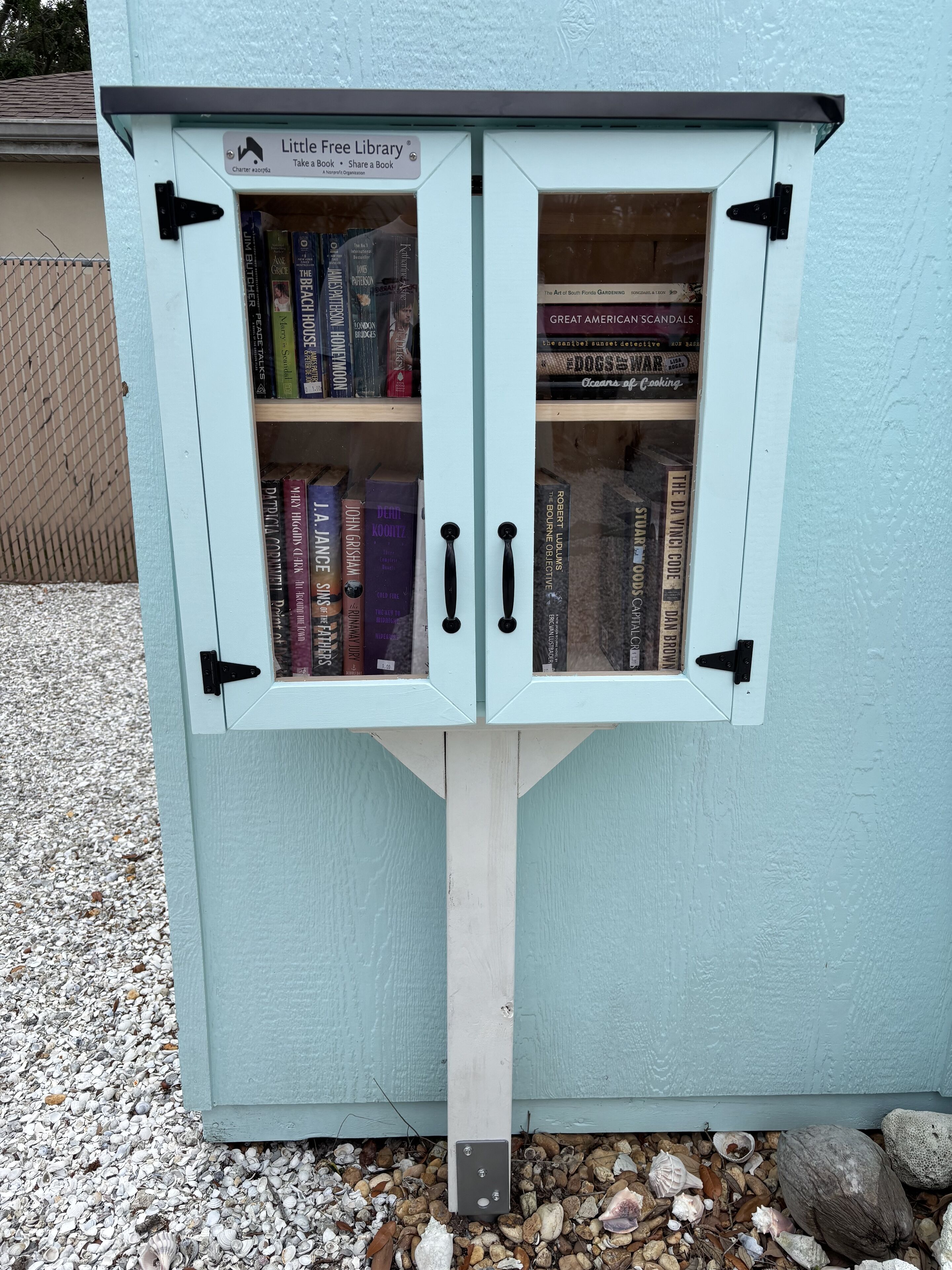 A Little Free Library with a white exterior and a wooden base, containing various books on shelves.