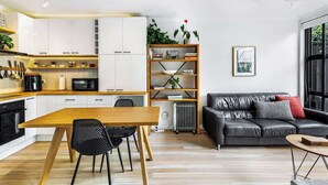 Una cocina moderna con armarios blancos, una mesa de comedor de madera y sillas negras. Hay un sofá de cuero negro y una estantería con plantas y libros.