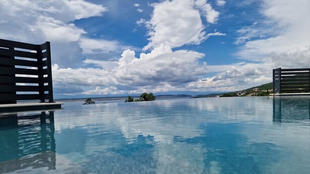 An infinity pool with clear blue water reflecting the sky and clouds, surrounded by wooden deck structures. - Villa V (Crikvenica)