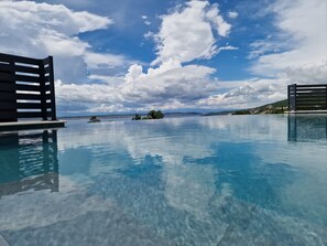Una piscina infinita con agua azul cristalina que refleja el cielo y las nubes, rodeada de estructuras de terraza de madera.