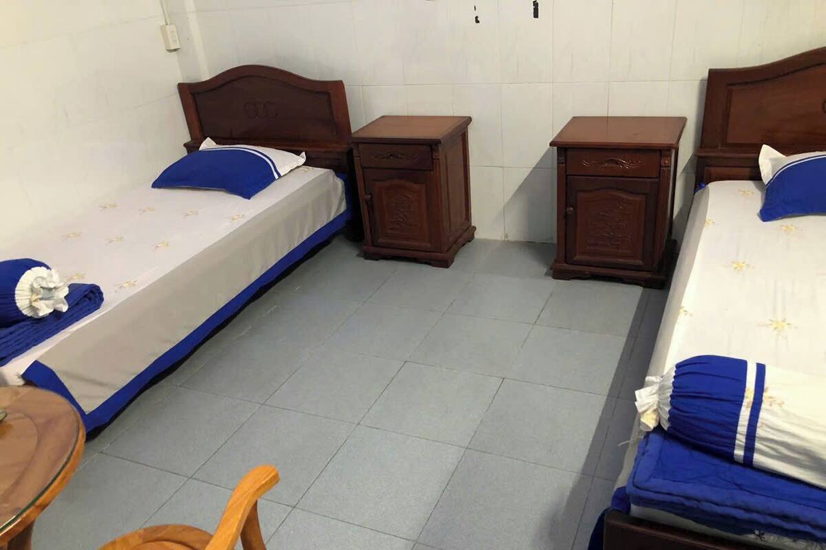 Ein Zimmer mit zwei Einzelbetten, jeweils mit einem Holz-Kopfteil und Nachttischen. Im Vordergrund steht ein kleiner Holztisch mit einem Stuhl.