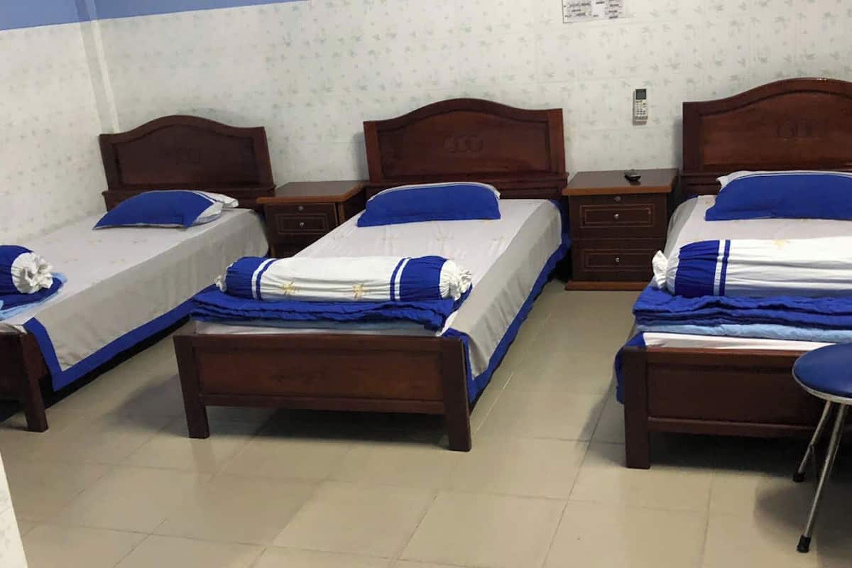 Ein Hotelzimmer mit drei Holzbetten, jeweils mit blauer und weißer Bettdecke und Kissen. Auf der rechten Seite der Betten befindet sich ein kleiner Nachttisch mit Telefon und Lampe.