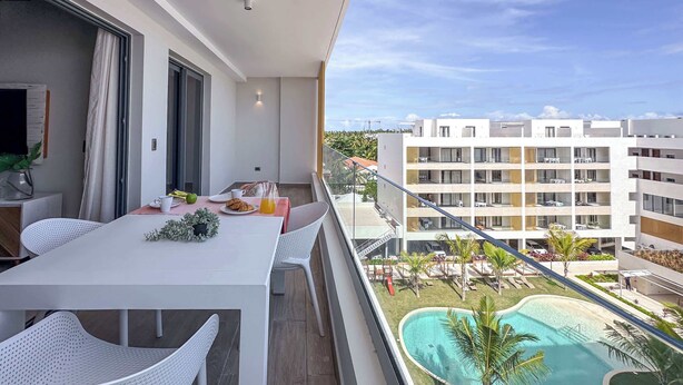 A balcony with a dining table, chairs, and a view of a swimming pool and buildings. - Gorgeous Penthouse with Rooftop and Ocean Views (Punta Cana)