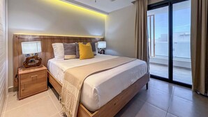 A modern hotel room with a large bed, wooden headboard, bedside tables with lamps, a ceiling fan, and a view of the city through large windows. - Gorgeous Penthouse with Rooftop and Ocean Views (Punta Cana)