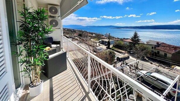 A balcony with a potted plant, a view of a coastal town, and a clear sky.