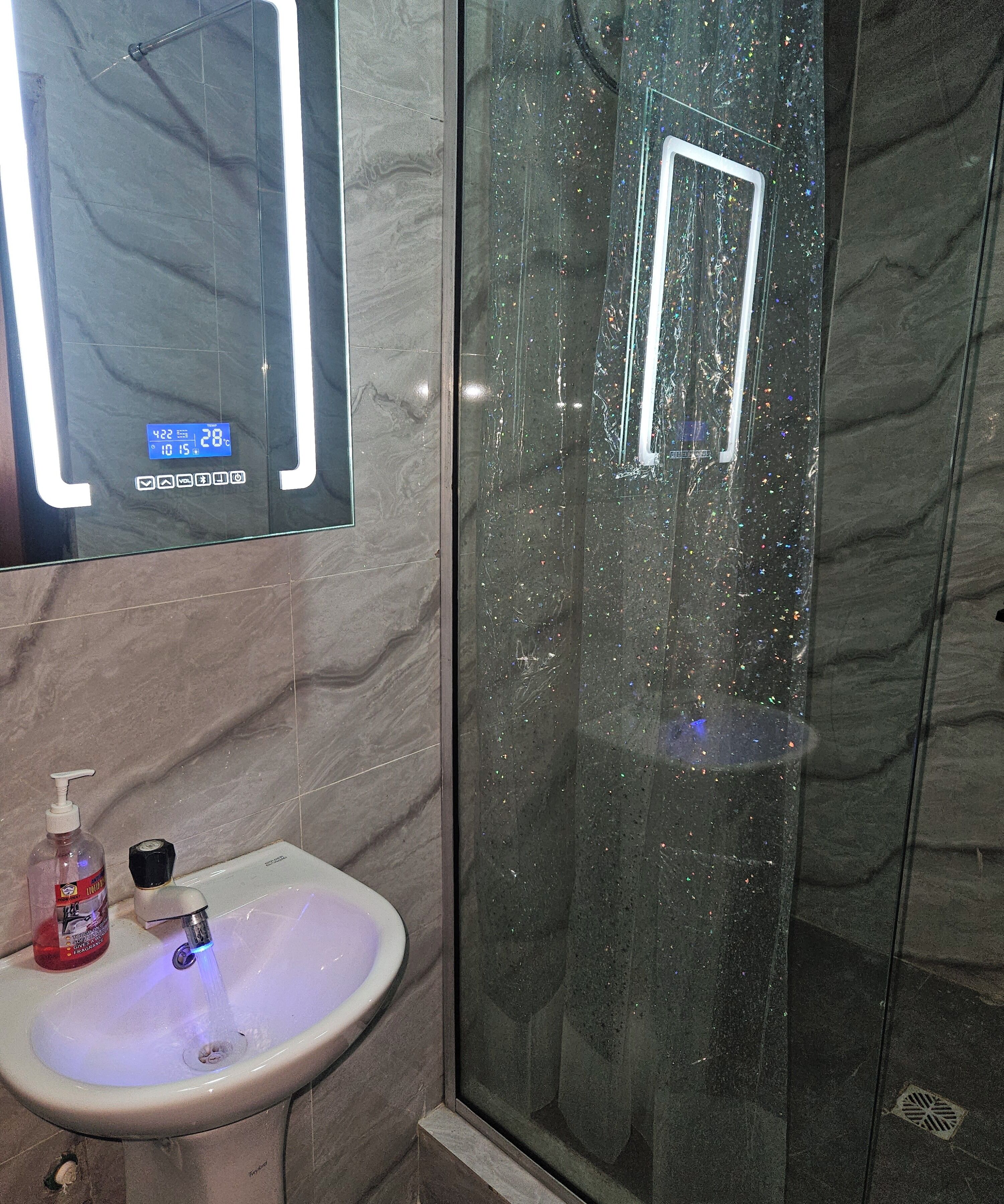 A bathroom with a glass-enclosed shower, a sink with a soap dispenser, and a digital thermometer displaying 28°C.