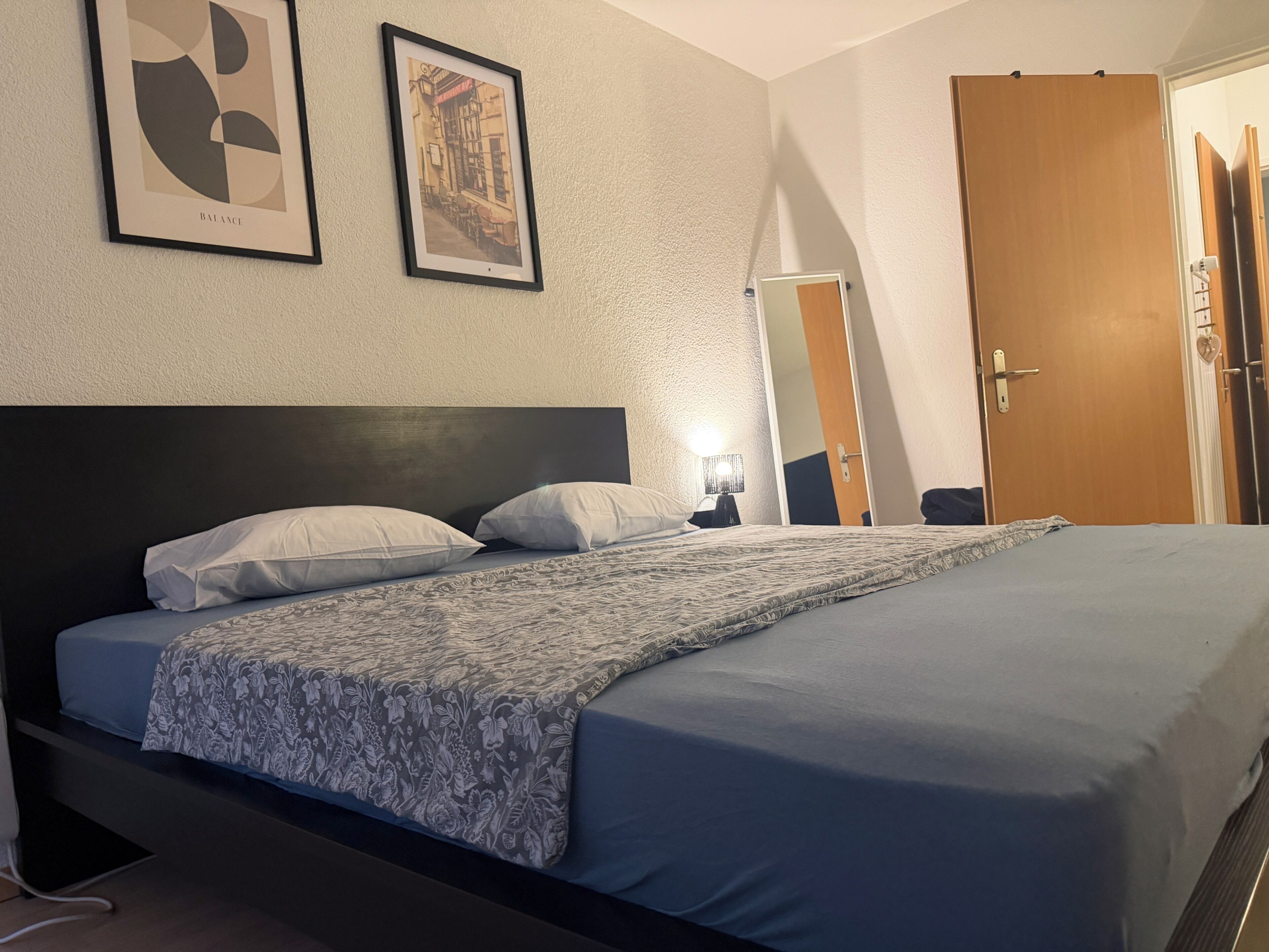 Een hotelkamer met een bed, nachtkastje, lamp, spiegel en een deur.