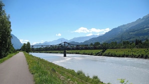 Un pont enjambant une rivière, avec un sentier qui longe la rivière, le tout entouré de montagnes et de verdure.