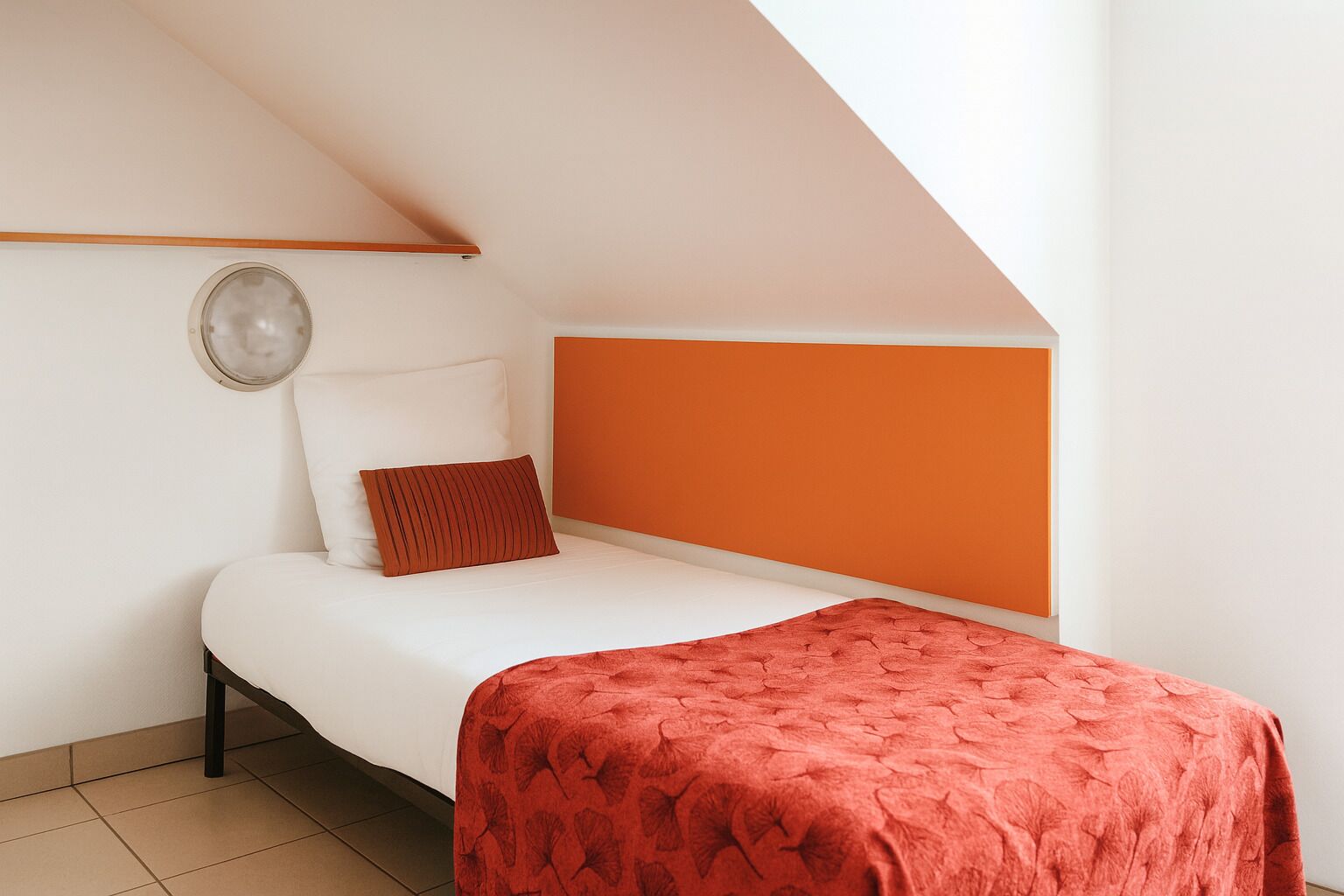 Une petite chambre avec un lit simple, un plafond en pente, des murs blancs et un mur d’accent orange.
