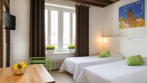 Ein Hotelzimmer mit zwei Betten, einem Holztisch mit Obstschale, einem grünen Sessel, einem Fenster mit Vorhängen und einem Bild an der Wand.