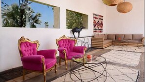 Ein Wohnzimmer mit zwei rosafarbenen Sesseln, einem Sofa, einem Glastisch und Blick auf Palmen durch große Fenster.