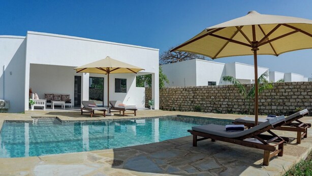 Un espace piscine avec des chaises longues et des parasols, un mur en pierre et un bâtiment blanc doté d’une porte et de fenêtres.
