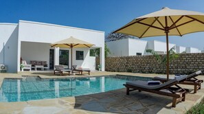 Un espace piscine avec des chaises longues et des parasols, un mur en pierre et un bâtiment blanc doté d’une porte et de fenêtres.