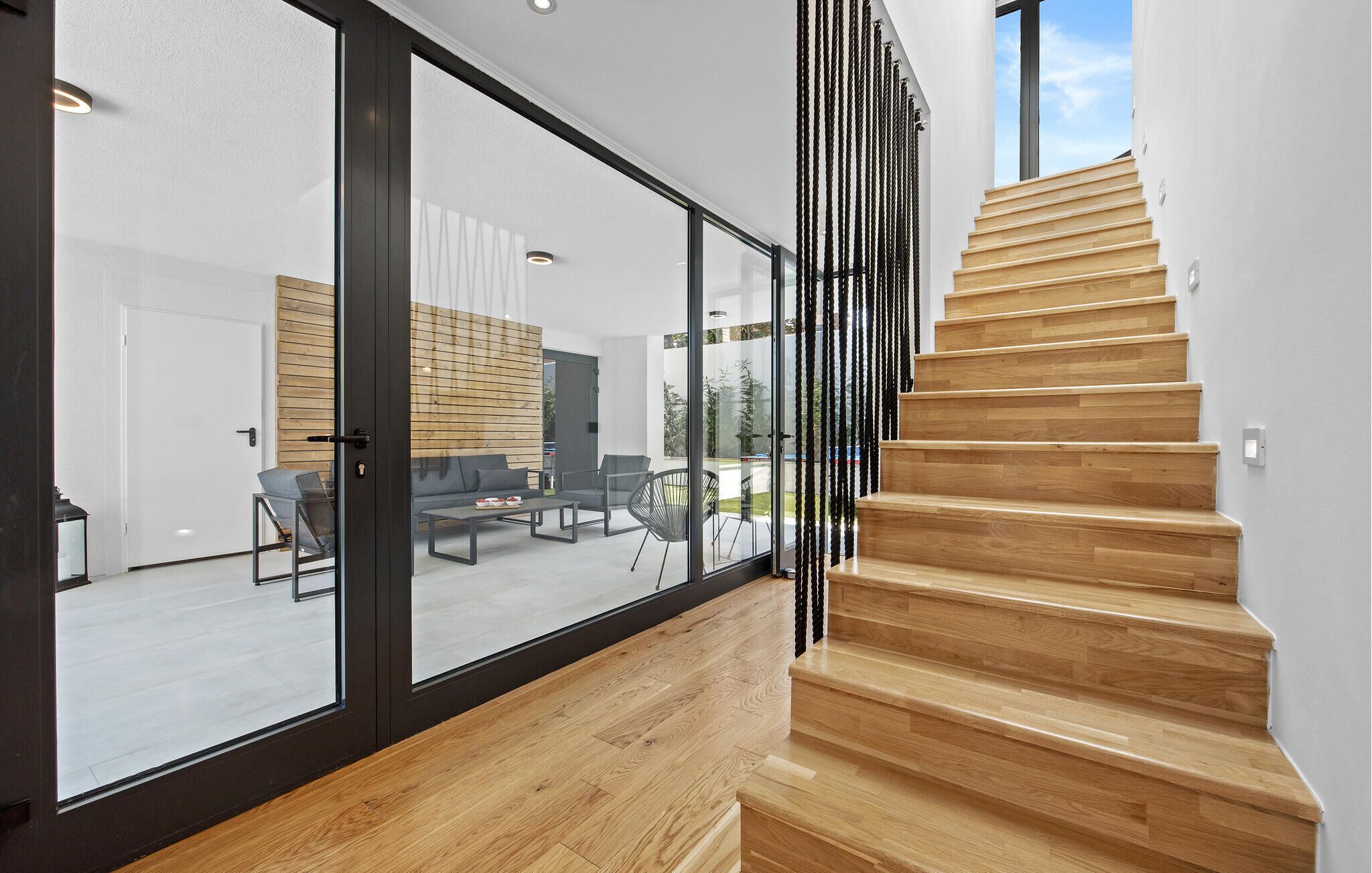 A modern interior with a wooden staircase, large glass doors leading to a living area with furniture and a view of the outdoors.
