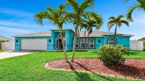 A two-story house with a turquoise exterior, a white garage door, and a well-maintained lawn with palm trees.