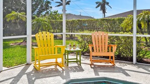 Two Adirondack chairs with a small table and two bottles of beer in between them, set on a patio with a view of a garden and a pool.