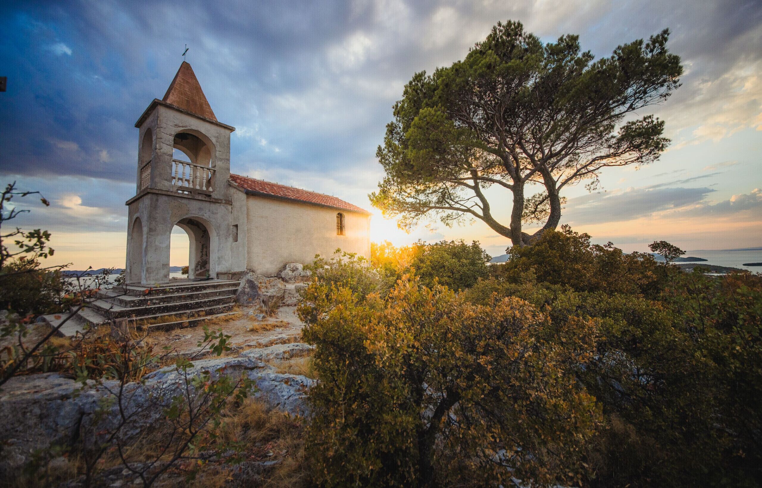 A church with a bell tower, surrounded by trees and a view of the sea.