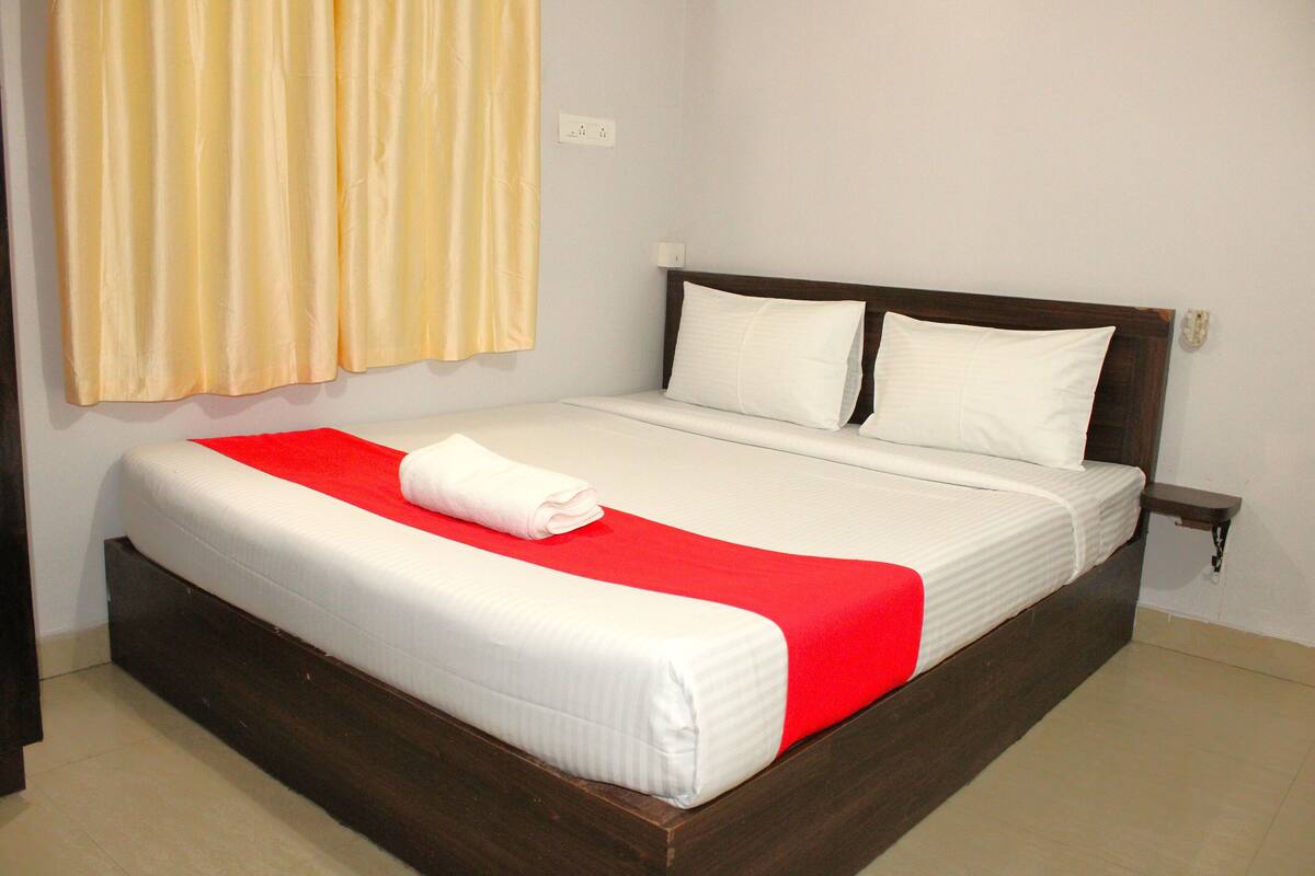 A neatly made bed with a red blanket and white pillows, a wooden headboard, and a bedside table with a power outlet.
