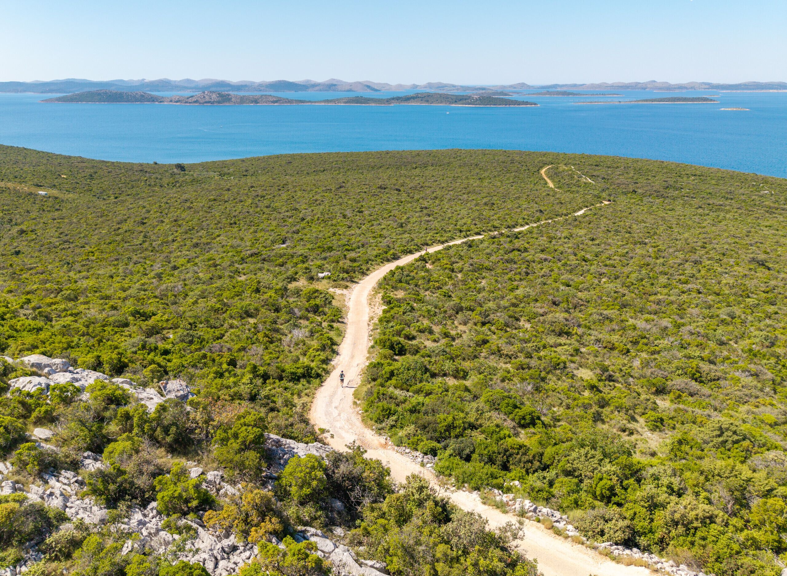 Ein unbefestigter Weg schlängelt sich durch ein hügeliges Gebiet mit dichter Vegetation und bietet einen Blick auf ein großes Gewässer in der Ferne.
