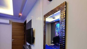 A hotel room with a wooden floor, a TV mounted on the wall, a large mirror with a decorative frame, and a bed with a blue bedspread. - VINANDA (New Delhi)