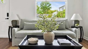 Un salon avec un canapé gris, une table basse noire ornée d’un vase et d’un bol, et une fenêtre donnant sur un paysage verdoyant et un immeuble.