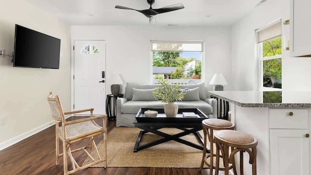 Un salon moderne avec un canapé, une table basse et deux tabourets de bar. On peut voir un ventilateur de plafond, et une fenêtre laisse entrer la lumière naturelle.