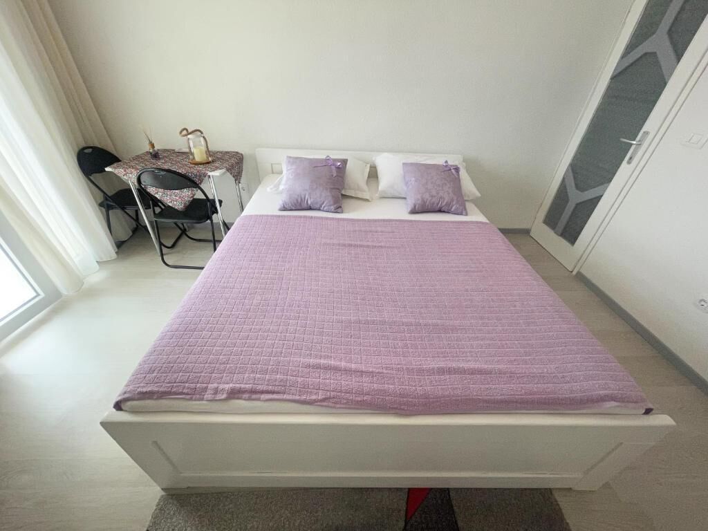 A neatly made bed with a purple bedspread and pillows, a small table with a lamp, and a chair in a room with a large window and a door with a geometric pattern.