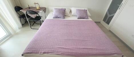 A neatly made bed with a purple bedspread and pillows, a small table with a lamp, and a chair in a room with a large window and a door with a geometric pattern.