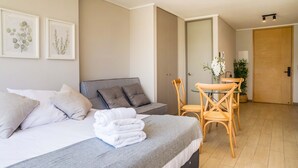 A neatly made bed with pillows, a grey blanket, and a folded white towel. There is a grey sofa with cushions, a wooden chair, and a small table with a vase of flowers. The room has a wooden floor and a door in the background. - Perfect and Cozy Studio in Nunoa Ideal for 2 (Ñuñoa)