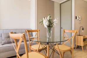 A modern living room with a glass-top dining table, two wooden chairs, a grey sofa, and a vase with white flowers.