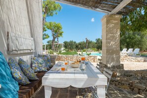 A white table set outdoors with a stone wall backdrop, a stone bench with patterned cushions, and a view of a swimming pool and trees.
