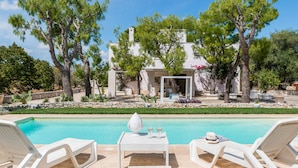 A poolside area with lounge chairs, a table with a vase and glasses, and a building with a white facade and trees in the background. - Villa Thea - Charming House (Carovigno)