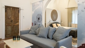 A room with a grey sofa, a bed with a round mirror, a wooden door, and a wall with a floral pattern. - Villa Thea - Charming House (Carovigno)
