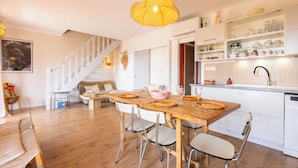 A spacious living area with a wooden dining table, chairs, a staircase, and a kitchen with shelves.