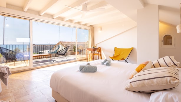 A bedroom with a large bed, a balcony with chairs, and a view of the sea.