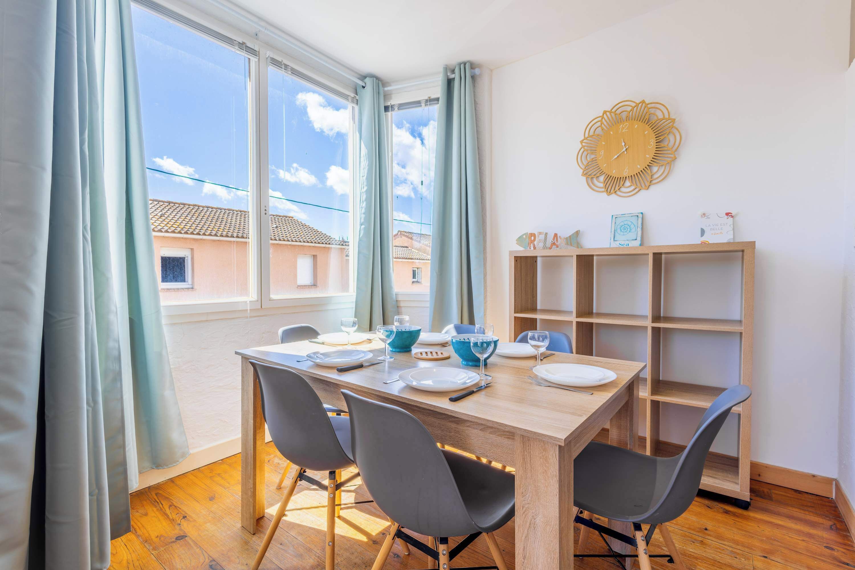 Un comedor con una mesa de madera, cuatro sillas, una estantería y una ventana con cortinas.